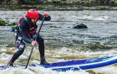 Individual in a red helmet paddleboarding on a river with white water.
