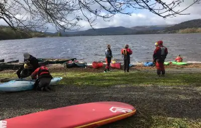 Group of people preparing kayaks and canoes by the shore of a lake on a cloudy day.
