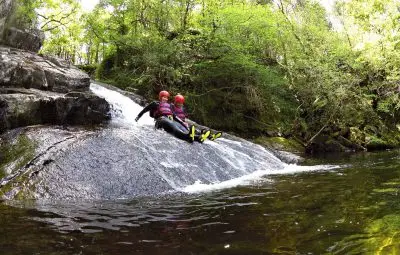 Two people in wetsuits sitting on a rock next to a small waterfall in a forested area.