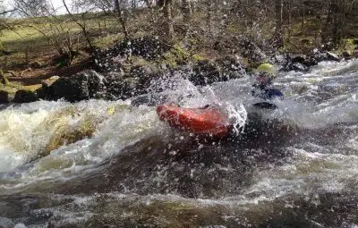 A kayaker in an orange kayak navigating through white water rapids with splashes of water.