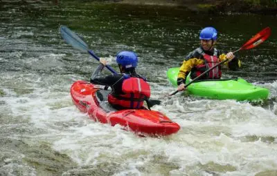 Two individuals in kayaks, one red and one green, paddling in a river with white water.