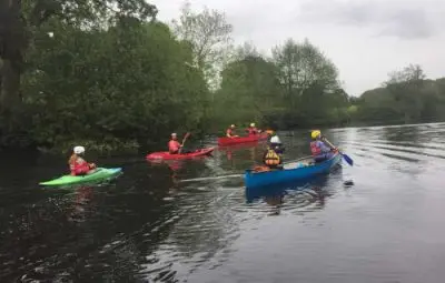 A group of six kayakers paddling on a calm river surrounded by greenery.