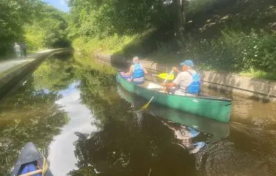 Two individuals paddling a green canoe on the Llangollen Canal surrounded by greenery.