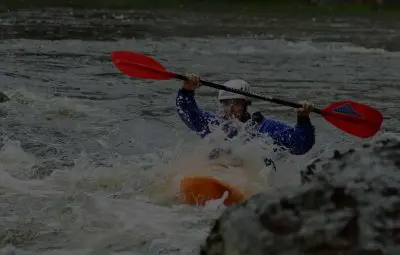 Individual paddling a kayak with red paddles in turbulent water surrounded by rocks.