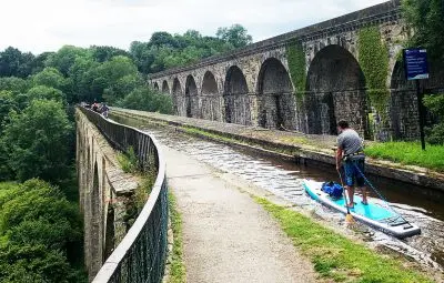 A person paddleboarding on the Llangollen Canal near Chirk Aqueduct with stone arches in the background.