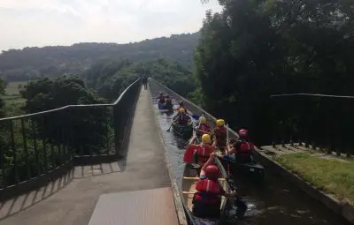 A group of people in canoes paddling along a canal under a bridge.