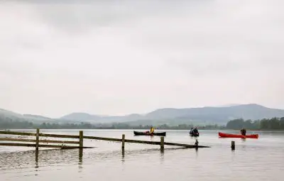 Two canoes, one red and one blue, on a lake with a wooden fence and mountains in the background.